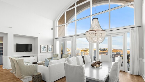 A spacious living room with a high ceiling, large windows, a chandelier, and a dining area with white furniture. - Gulf-Front Retreat with Panoramic Views (Watersound)