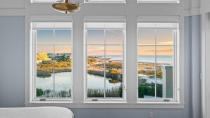 A bedroom with three large windows offering a view of a coastal landscape with buildings and a body of water. - Gulf-Front Retreat with Panoramic Views (Watersound)