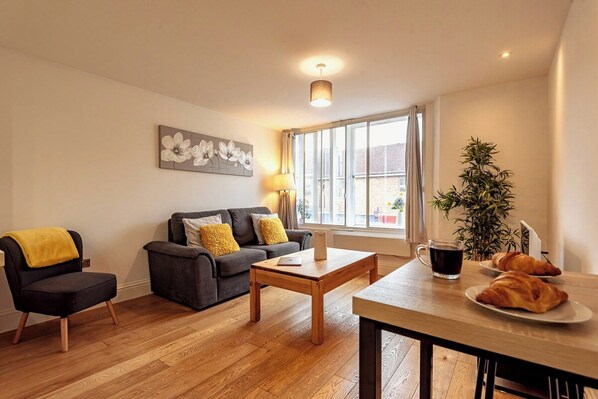 A living room with a sofa, armchair, coffee table, and a dining table with croissants and a coffee mug.