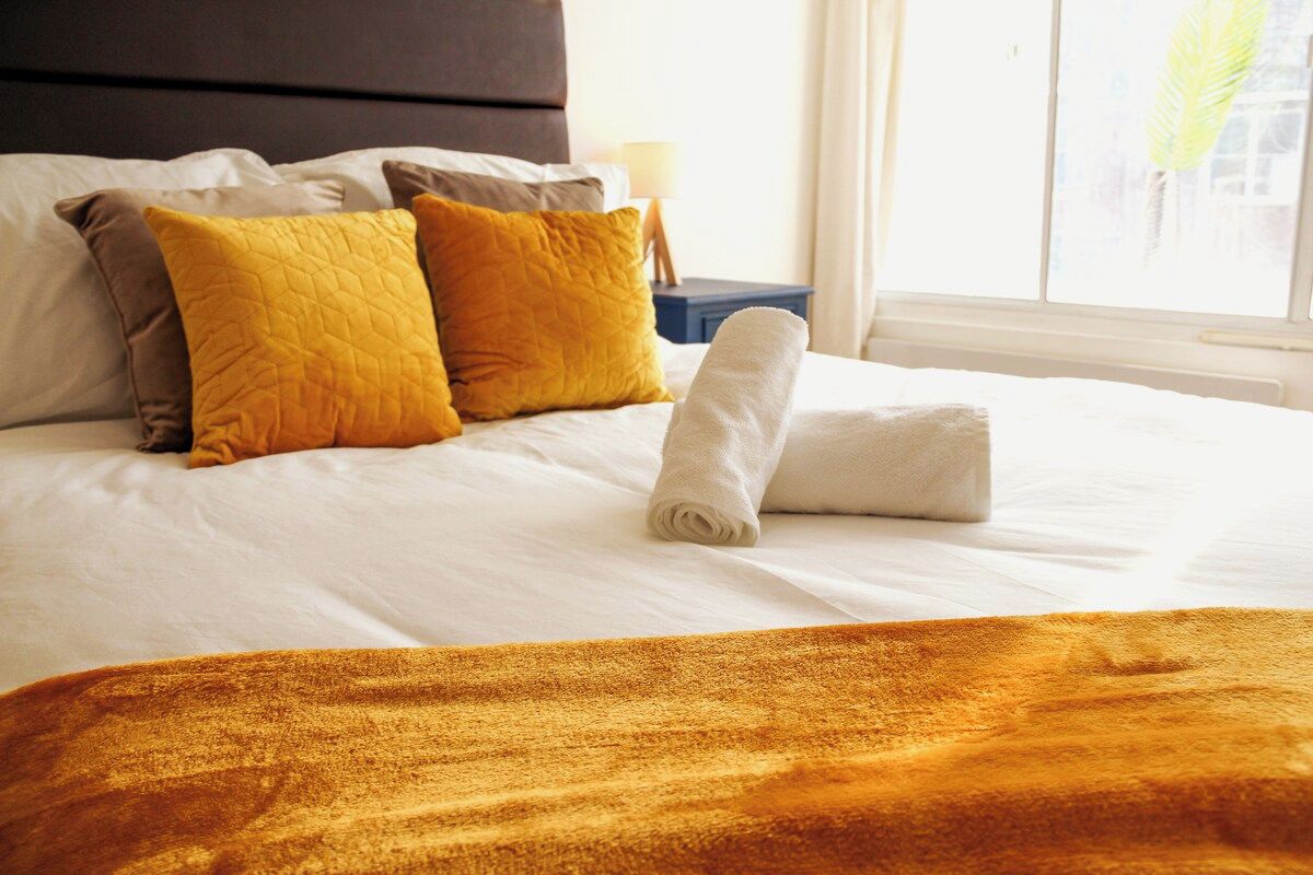 A neatly made bed with a rolled white towel, two pillows in shades of yellow and brown, and a brown blanket.