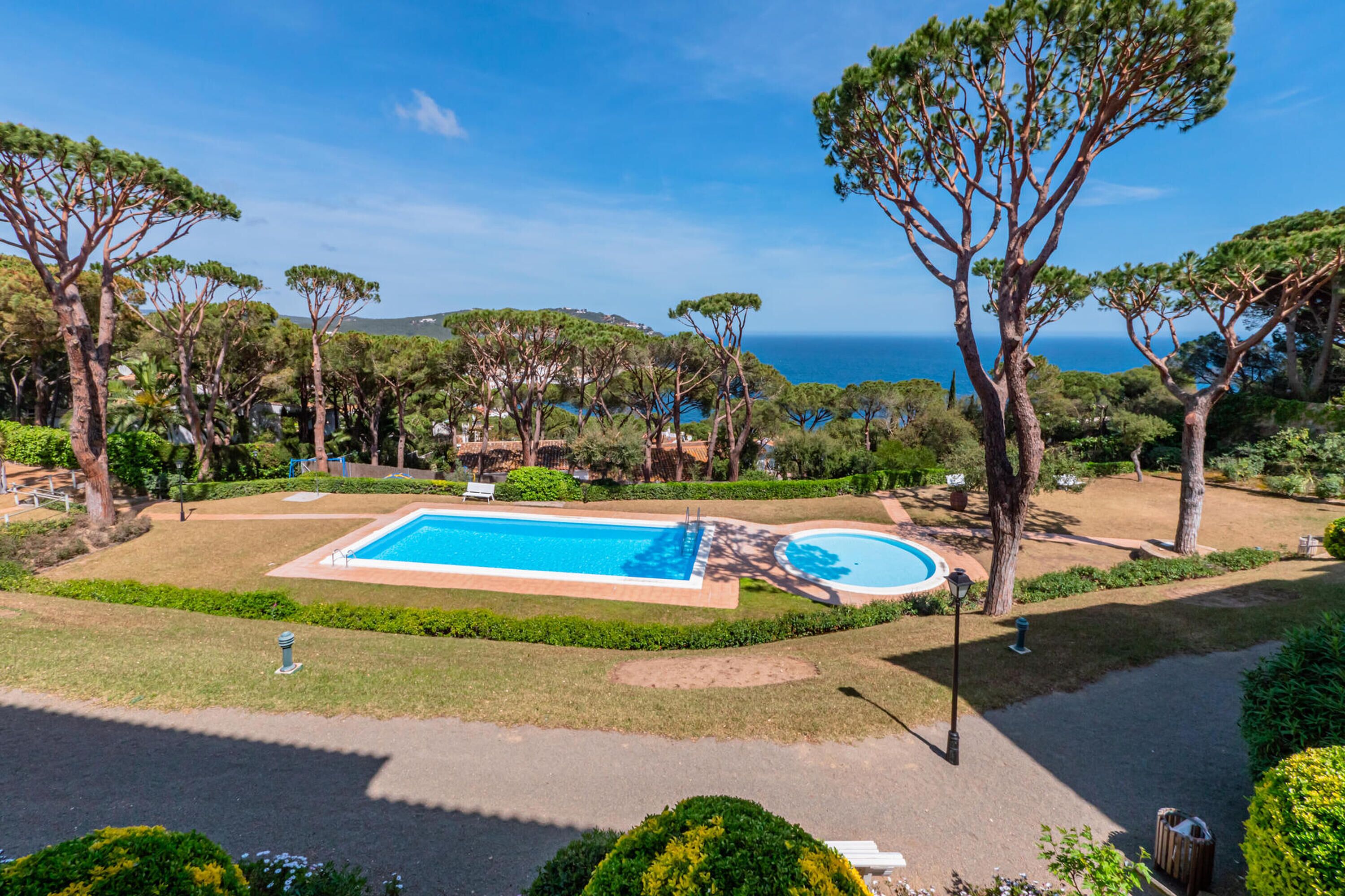 A pool area with a rectangular pool and a smaller circular pool, surrounded by trees and a view of the sea.