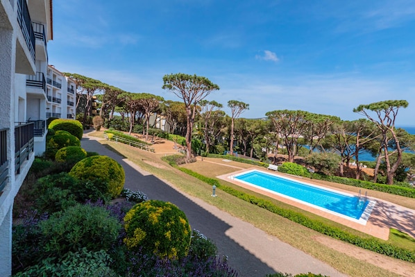 A pool area with a clear view of the sea, surrounded by well-maintained greenery and a pathway leading to a building with balconies.