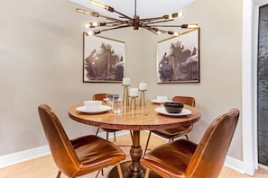 A round dining table with six brown leather chairs, a wooden table top, and a modern chandelier with exposed bulbs. Two framed abstract paintings hang on the wall above the table.