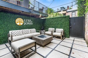 Un espace patio aménagé avec un canapé d’angle, deux fauteuils et une table basse. On aperçoit un arbuste et un portail à l’arrière-plan.