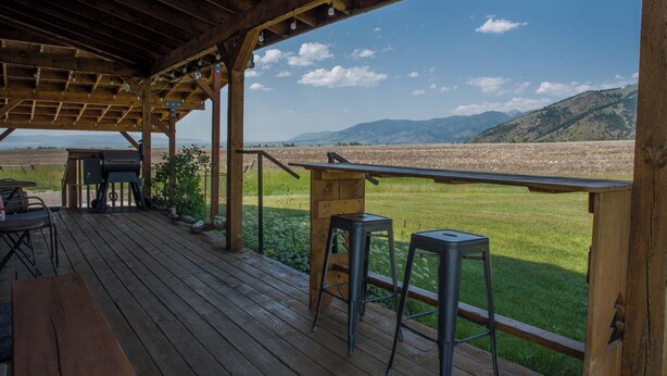 A wooden deck with a table and chairs, overlooking a grassy field and distant mountains. - Cozy Cabin, Mountain View, AC, Porch, Fireplace, Wildlife, 4 mi to town. (Bozeman)