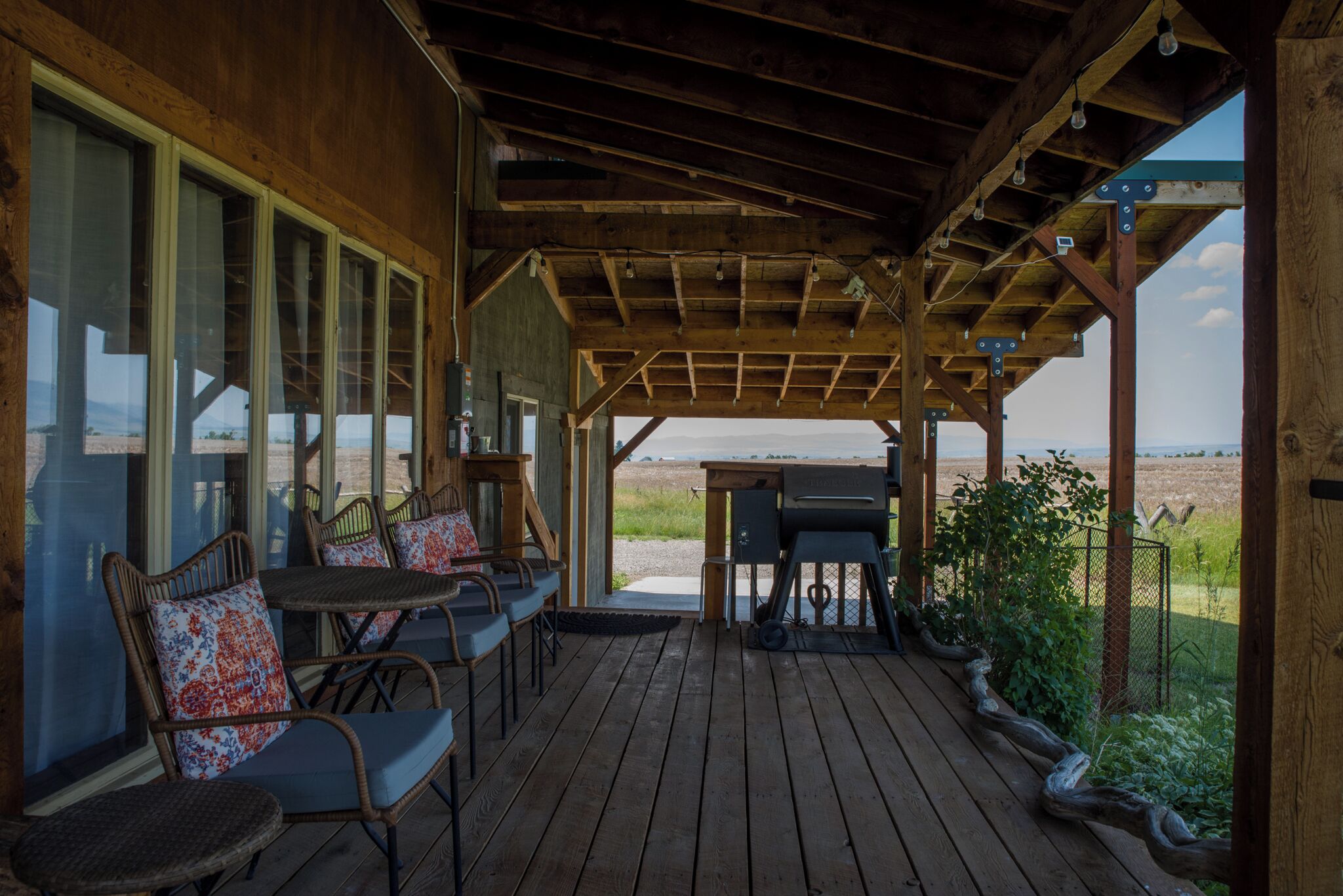 A covered outdoor seating area with wooden tables and chairs, a grill, and a view of the landscape.