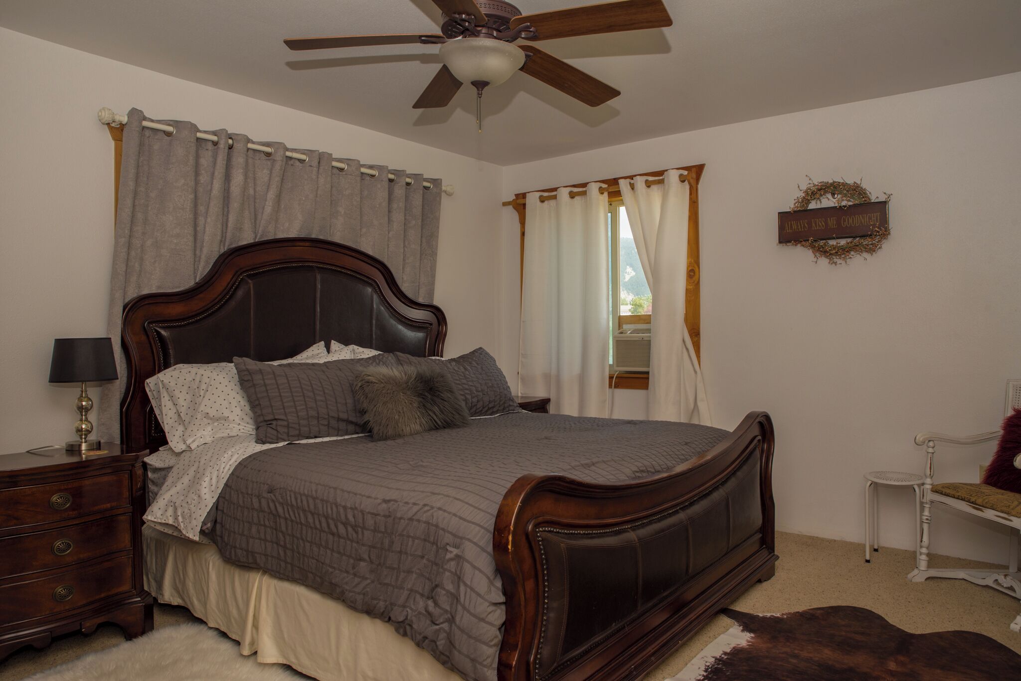 Quarto com cama de madeira, mesa de cabeceira, luminária, ventoinha no teto e janela com cortinas.