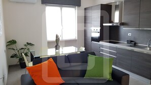 Een moderne woonkamer met een bank en twee kussens, een glazen salontafel, een potplant en een raam met zonwering.