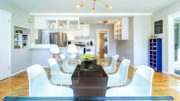 Une cuisine moderne avec des meubles blancs, un réfrigérateur en acier inoxydable et un coin repas comprenant une table en verre et des chaises blanches.
