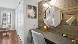 A hotel room with a wooden wall, a round mirror, a desk, a chair, and a window with a view.
