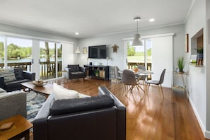 A spacious living room with a large sofa, a coffee table, and a dining area with chairs and a table. There is a TV mounted on the wall and a plant on a stand. - Renovated, Lakefront, Pinehurst #5, Ground Level, 2BR, 2BA, Pool, WiFi, Cable (Pinehurst)