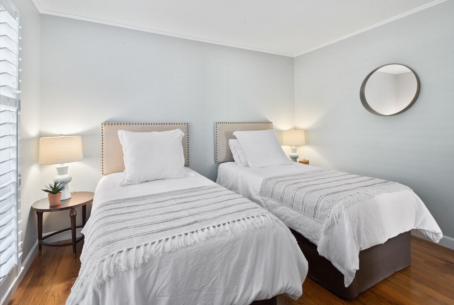 A room with two beds, each with a headboard, bedside tables, and lamps. There is a round mirror on the wall.