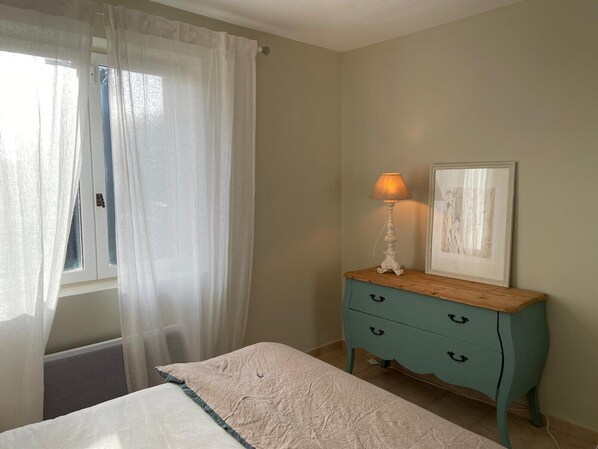 A bedroom with a bed, a nightstand with a lamp, a framed picture, and a window with sheer curtains. - Charming farmhouse in peaceful Provence, between the Luberon and the Alpilles (Robion)