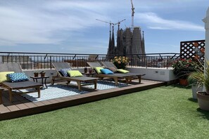 Terraço no telhado com espreguiçadeiras e grama sintética, com vista para um canteiro de obras e uma catedral.