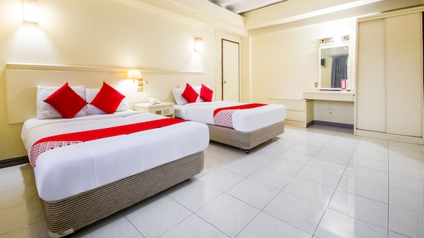 A hotel room with two beds, each with red pillows, a nightstand, a mirror, and a bathroom with a sink and mirror.