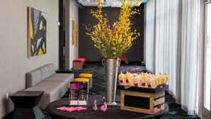 A modern hotel lobby with a seating area, a decorative vase with yellow flowers, and a display of snacks. - Aloft by Marriott Dallas DFW Airport Grapevine (Coppell)