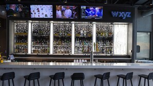 A bar with multiple screens displaying content, a well-stocked shelf with various bottles, and a bar counter with stools. - Aloft by Marriott Dallas DFW Airport Grapevine (Coppell)