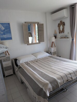 A bedroom with a bed, bedside tables, a mirror, an air conditioning unit, and a window with shutters.