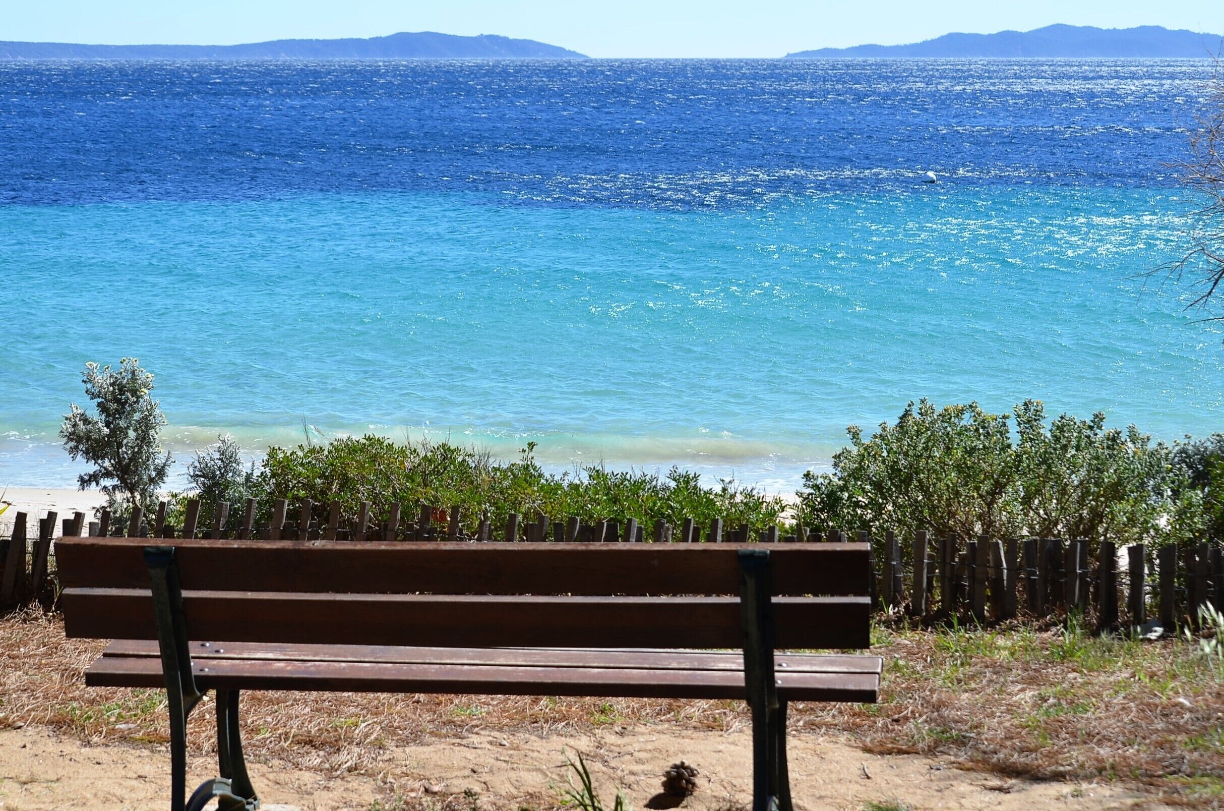 Eine Bank mit Blick auf einen Strand, klares blaues Wasser und ferne Berge.