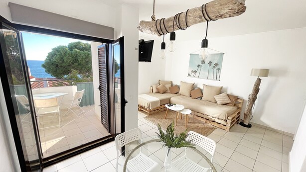 En moderne stue med sofa, et glasbord og stole, en balkon med havudsigt og en rustik lysekrone i træ.