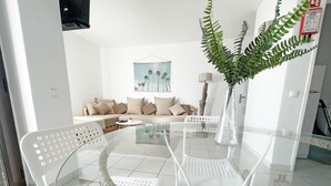 En moderne stue med et glasbord, hvide stole, en sofa med beige puder og et bregnehoved i en vase.