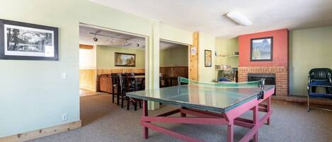 A room with a ping pong table, chairs, a fireplace, and framed pictures on the walls.