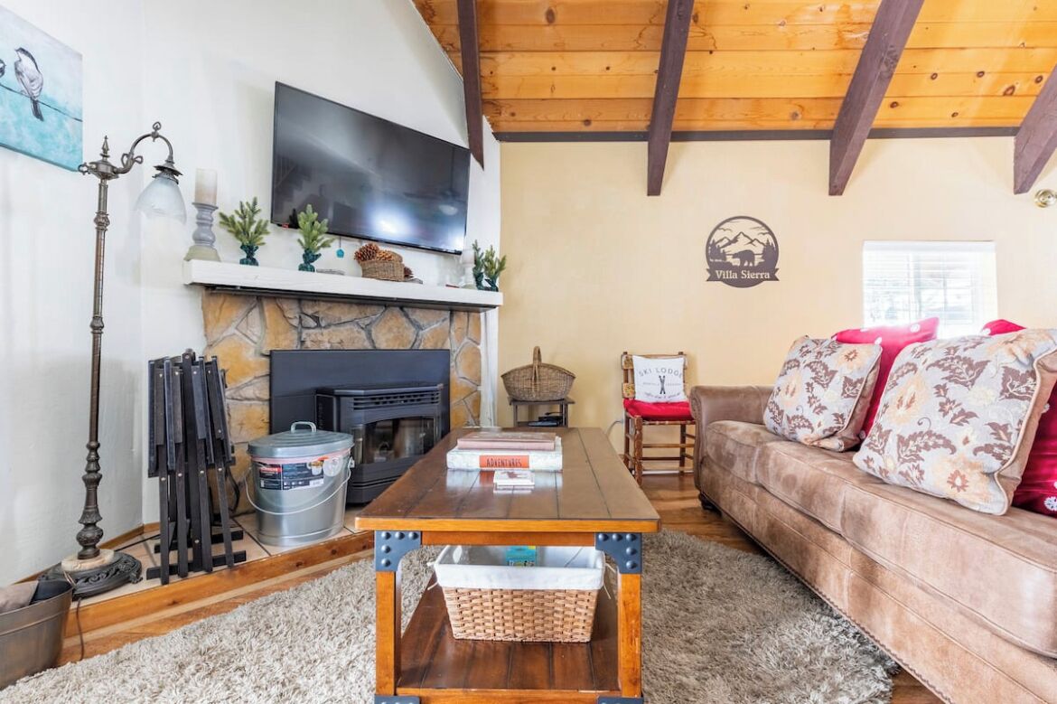 A cozy living room with a stone fireplace, a flat-screen TV, a wooden ceiling, a sofa with patterned cushions, and a sign indicating 'White Sierra'.