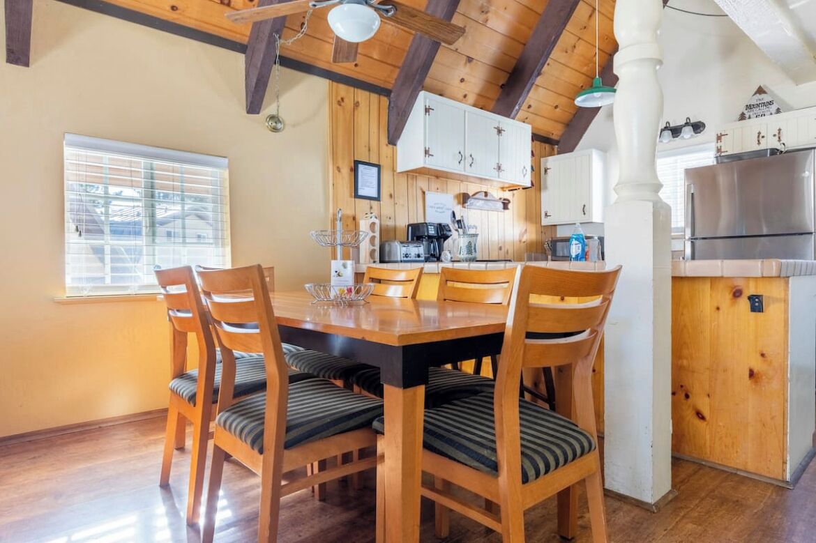 A wooden dining table with six chairs, a kitchen area with white cabinets, a refrigerator, and a window with blinds.