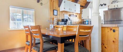 A wooden dining table with six chairs, a kitchen area with white cabinets, a refrigerator, and a window with blinds.