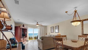 Uma sala de estar ampla com uma grande janela que oferece vista para o oceano, uma mesa de jantar com cadeiras e um ventilador de teto.