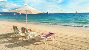 A beach with white lounge chairs and a table under a white umbrella, with the ocean and sailboats in the background. - Grand Blue Beach Hotel (Boracay Island)