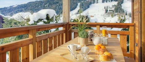 A wooden deck with a table set for breakfast, overlooking a snowy mountain landscape.