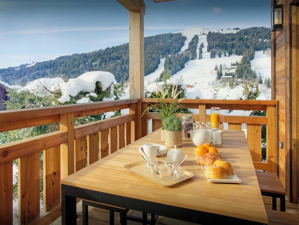 A wooden deck with a table set for breakfast, overlooking a snowy mountain landscape.