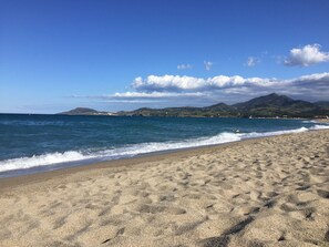 A sandy beach with gentle waves, a clear sky, and distant mountains. - Charming 60 m2 house with privacy in a quiet and safe wooded residential community (Argelès-sur-Mer)