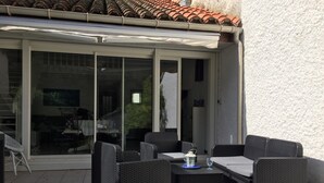 Een terras buiten met rieten meubels, een tafel en een glazen deur die naar de binnenruimte leidt.