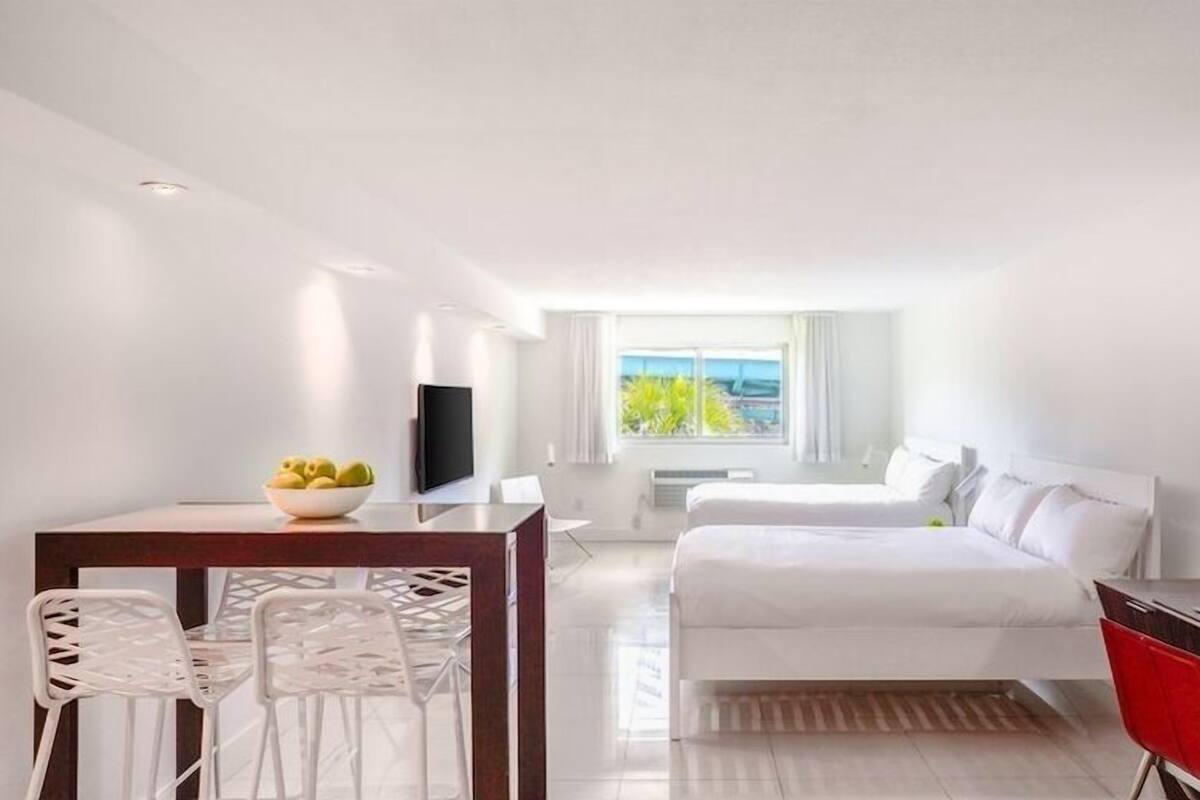 Quarto de hotel com cama, escrivaninha com laptop, área de refeições com cadeiras, televisão e janela com vista para o mar.