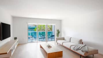 Una sala de estar moderna con televisor de pantalla plana, un sofá, una mesa de centro y un ventanal corredizo que da a un espacio exterior con sillas y plantas.