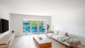 A modern living room with a flat-screen TV, a sofa, a coffee table, and a sliding glass door leading to an outdoor area with chairs and plants. - Beach Haus Bal Harbour (Bal Harbour)