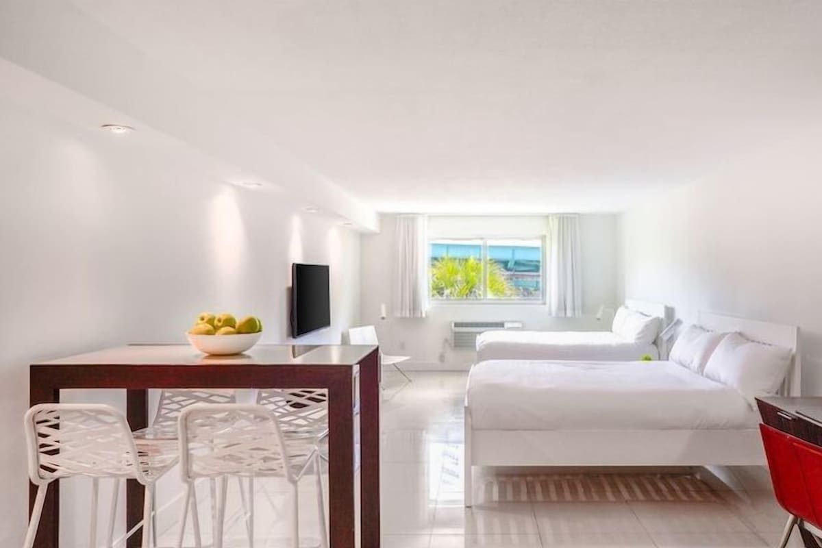A hotel room with a bed, a desk with a laptop, a dining area with chairs, a TV, and a window with a view of the sea.
