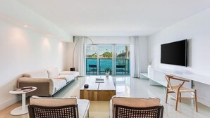 Un salon moderne avec un canapé, une table basse et des fauteuils. Un téléviseur est fixé au mur, et de grandes fenêtres offrent une vue sur la mer.