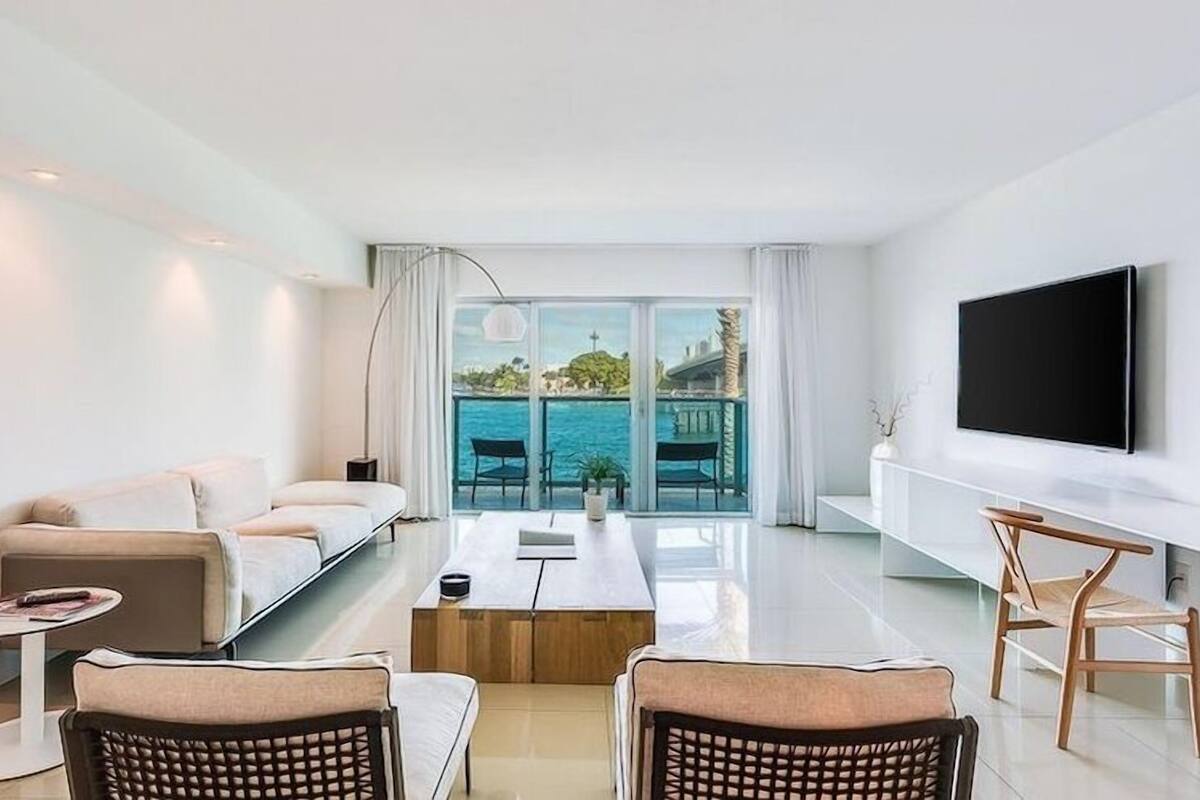 Uma sala de estar moderna com sofá, mesa de centro e cadeiras. Uma TV está instalada na parede, e há vista para o mar através de grandes janelas.