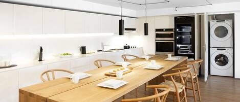 A modern kitchen with white cabinetry, a wooden dining table, and built-in appliances including a double oven and a stacked washer and dryer.