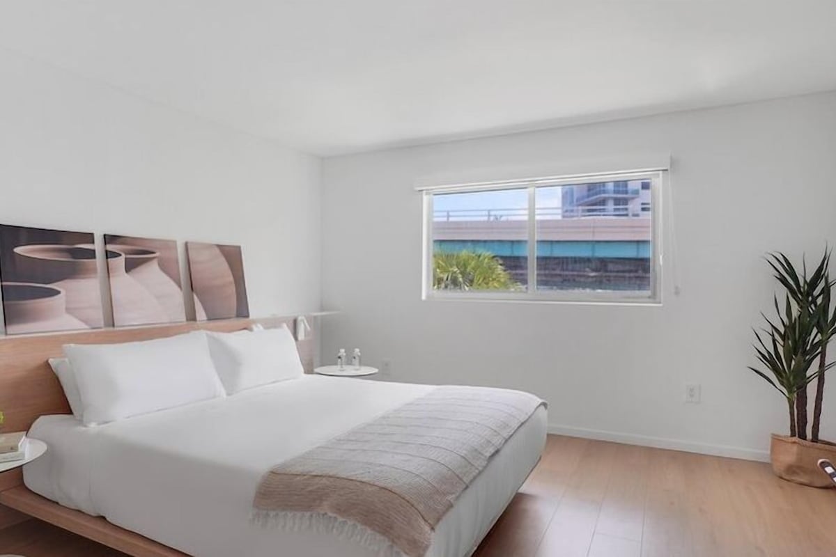 A modern hotel room with a large bed, a bedside table with a lamp, a small potted plant, and a window with a view of buildings.