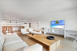 Ett modernt vardagsrum med en soffa, ett soffbord och stolar. Det finns ett köksområde med skåp och ett matbord. En platt-TV är monterad på väggen.