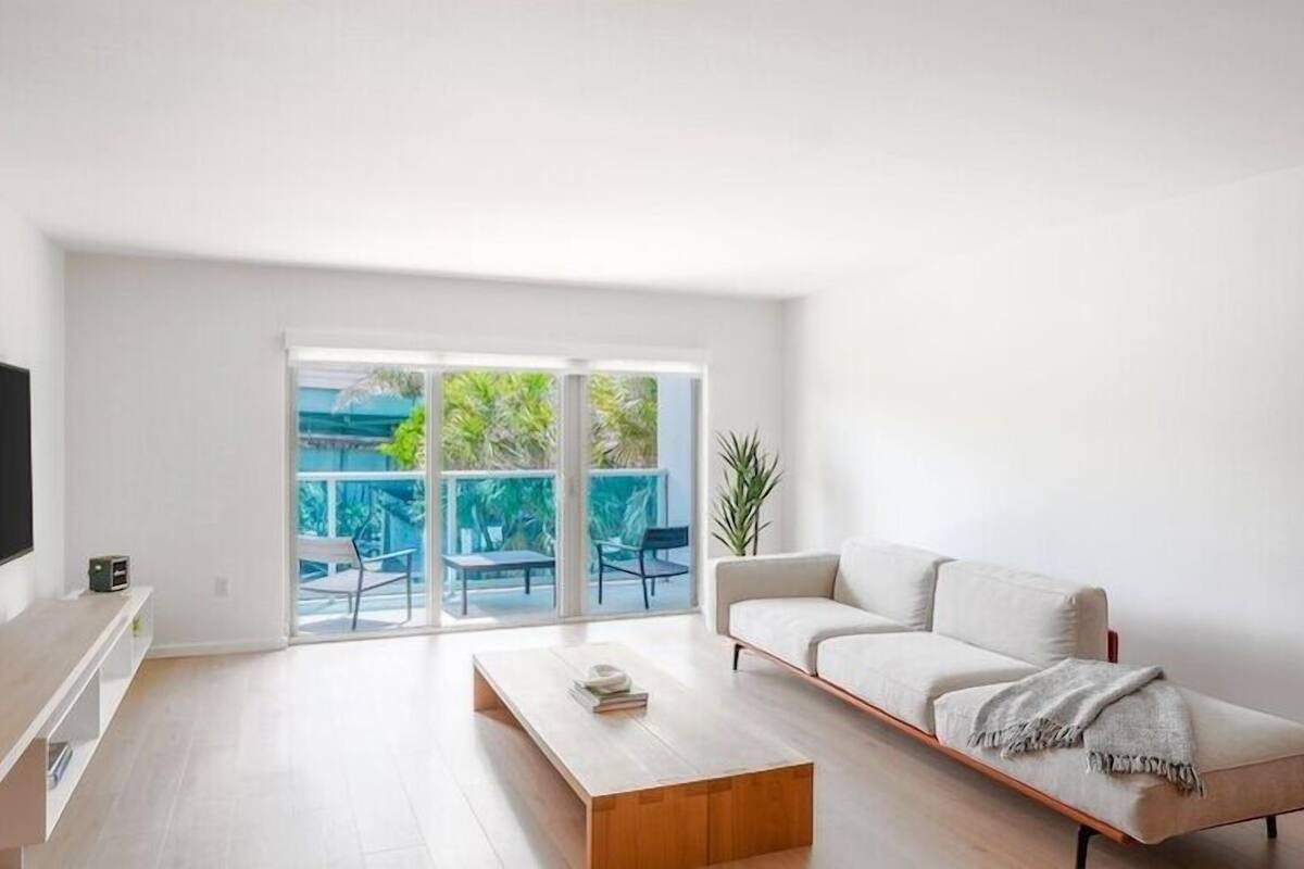 Uma sala de estar moderna com televisão de tela plana, sofá, mesa de centro e uma porta de vidro deslizante que dá para uma área externa com cadeiras e plantas.