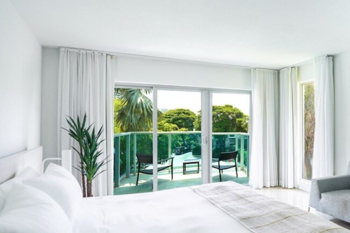 A modern hotel room with a large bed, a grey sofa, a flat-screen TV, and a balcony with a view of greenery.