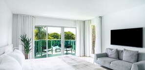 A modern hotel room with a large bed, a grey sofa, a flat-screen TV, and a balcony with a view of greenery. - Beach Haus Bal Harbour (Bal Harbour)