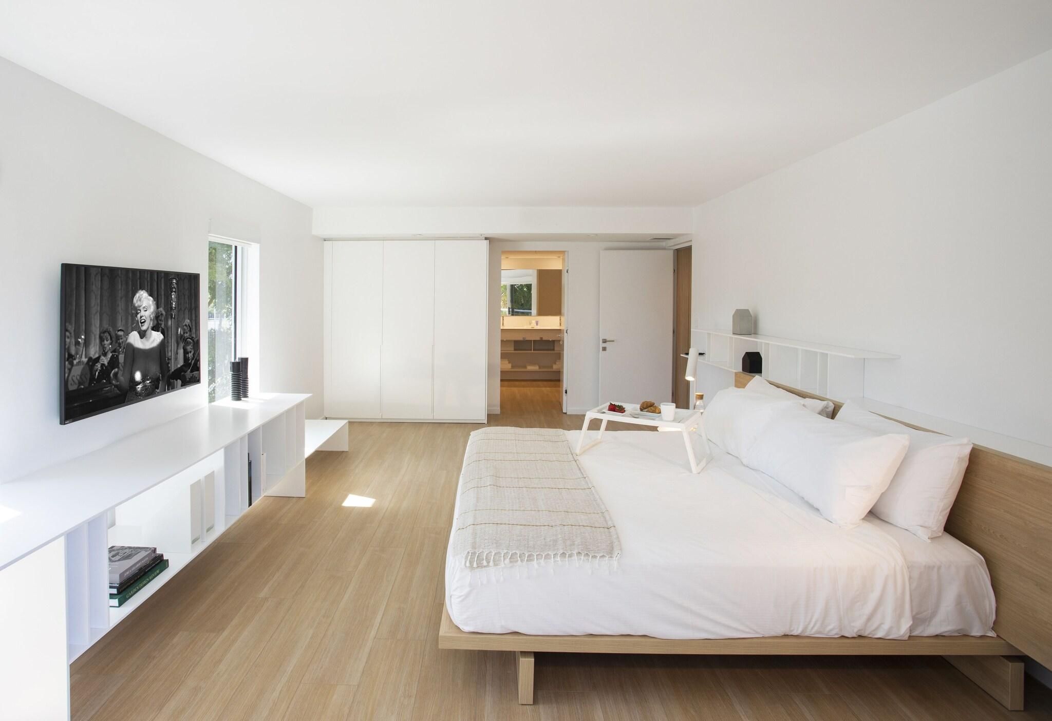 A modern bedroom with a large bed, a bedside table, a TV on the wall, and a staircase leading to an upper floor.
