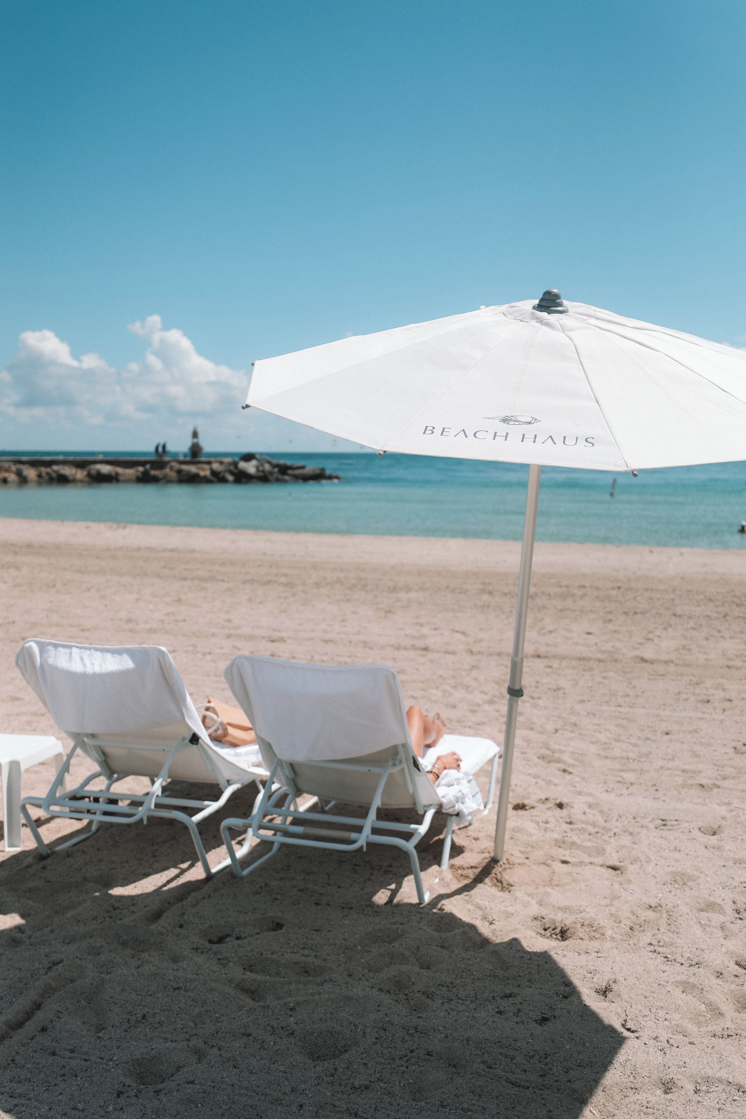 Duas cadeiras de praia sob um guarda-sol branco com a inscrição 'BEACH HOUSE', posicionadas em uma praia arenosa com céu claro e mar calmo ao fundo.
