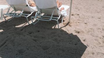 Duas cadeiras de praia sob um guarda-sol branco com a inscrição 'BEACH HOUSE', posicionadas em uma praia arenosa com céu claro e mar calmo ao fundo.